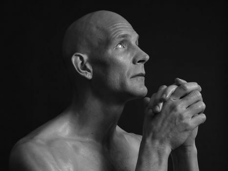 A bald shirtless man clasps his hands in prayer, eyes pleading skyward, over a black background. In black and white.