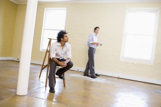 Man sitting on ladder in empty space with another man holding pa