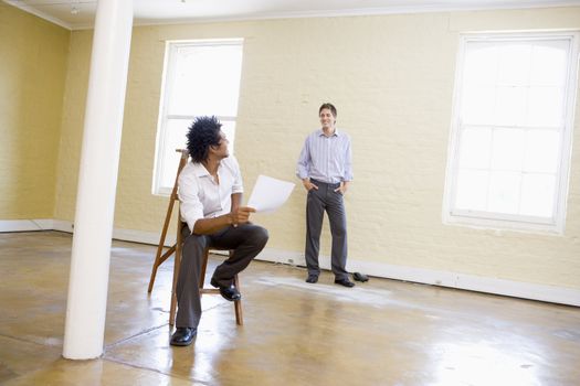 Man sitting on ladder in empty space holding paper talking to other