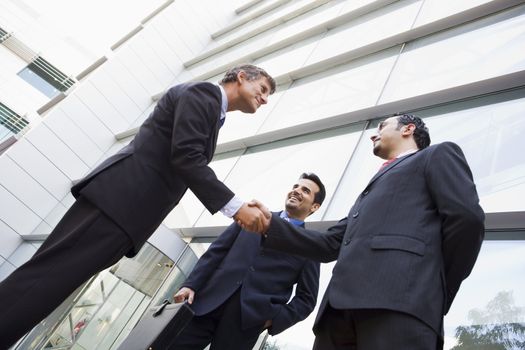 Three businessmen standing outdoors by building shaking hands and smiling (high key/selective focus) 