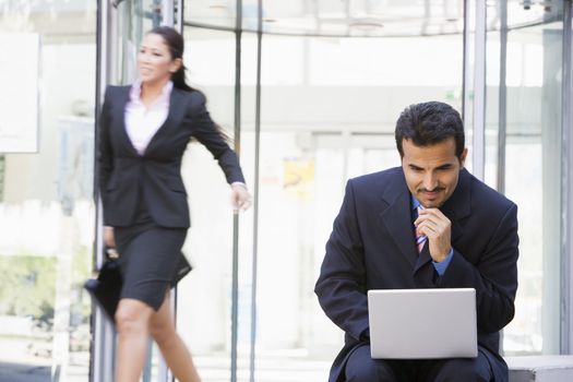 Businessman outdoors in front of building using laptop with businesswoman in background smiling (high key/blur) 