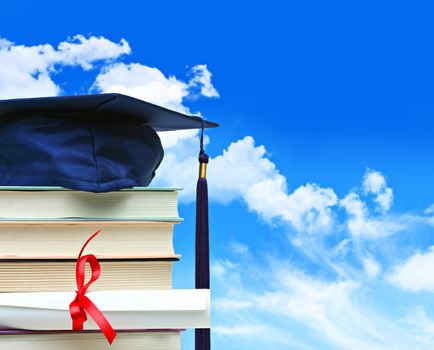 Stack of books with cap and diploma against blue sky