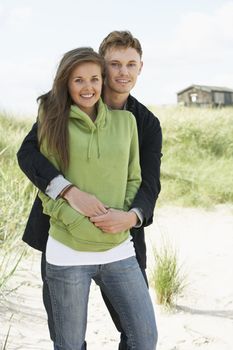Romantic Young Couple Standing By Dunes With Beach Hut In Distan