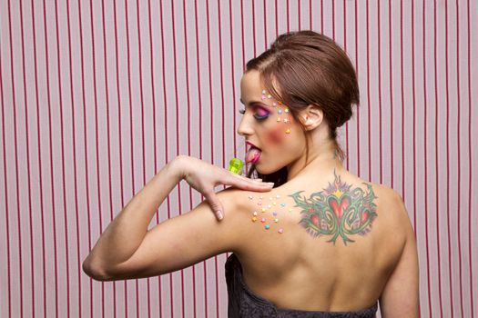 Young woman with colorful makeup and star candy glued to her face and body, licking a lollipop ring