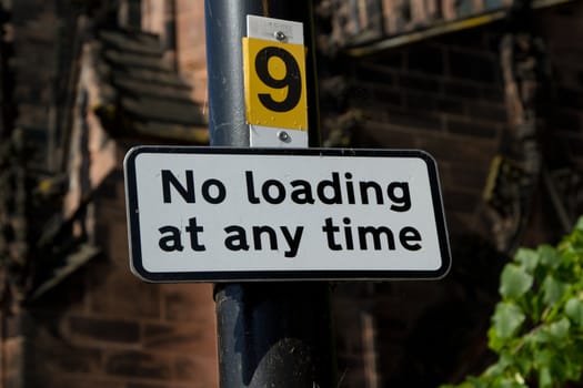A white rectangular sign with a black border and the words 'NO LOADING AT ANY TIME' attached to a post with a number '9'.