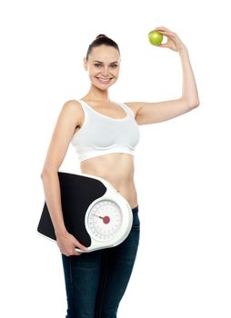 Attractive young female holding weighing machine and green apple standing against white background