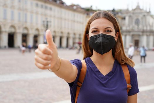 COVID-19 Optimistic traveler woman wearing black protective mask KN95 FFP2 showing thumbs up in city square