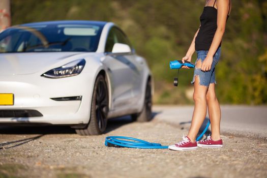 Beautiful young girl next to an electric car. Standing with a blue charging cable
