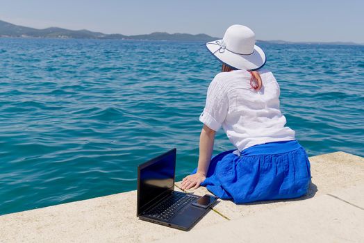 Work from anywhere. Side view of young woman, female freelancer in straw hat working on laptop while sitting on the beach. young woman working remotely on laptop by the sea