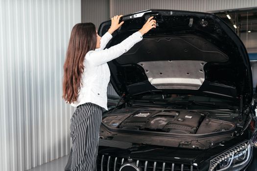 Young woman with opened hood in garage looking under car hood