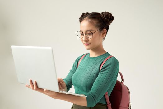 Charming clever lady is standing on the pure grey background with laptop in her hands, smiling, looking at it while browsing, typing, writing, wearing black specs