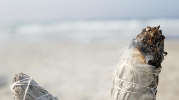 Dried white sage smoke, dry smudge stick burning, soft focus bokeh. Aroma smudging close up on ocean beach, aromatherapy meditation by sea water waves on sandy coast. Yoga, peace, harmony and ayurveda