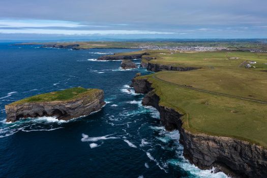 Aerial panoramic view of beautiful and green mountain of the famous tourist landmark Moher Cliff in Ireland with sea waves hitting rocks during day under cloudy sky