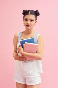 Photo of pretty student girl 20s with double buns hairstyle wearing  backpack holding many studying books isolated over pink background