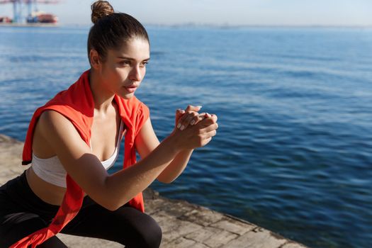 Close-up of focused young female athlete workout on a pier, doing squats and looking at sea