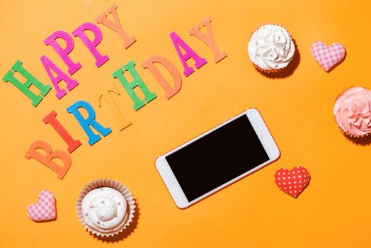 Words Happy Birthday with cupcake and smartphone on yellow background.