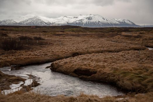Icelandic landscape with stream through grassy field with snow capped mountains in the distance atmospheric cloudy textured