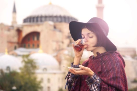 A young girl in a hat and poncho holds traditional Turkish tea