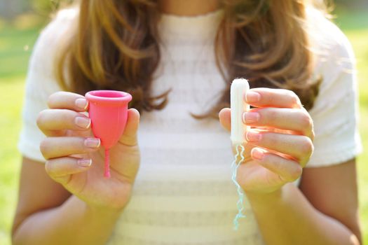 Young woman holding collector menstrual cup and tampon in her hands against green natural background