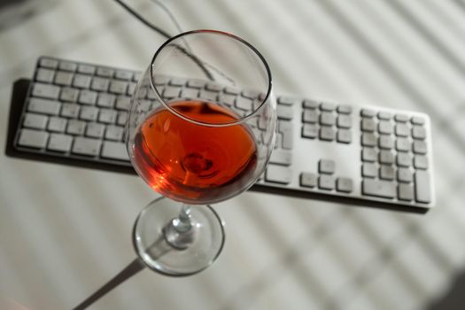 A goblet with red wine and a keyboard on a white table with shade from blinds