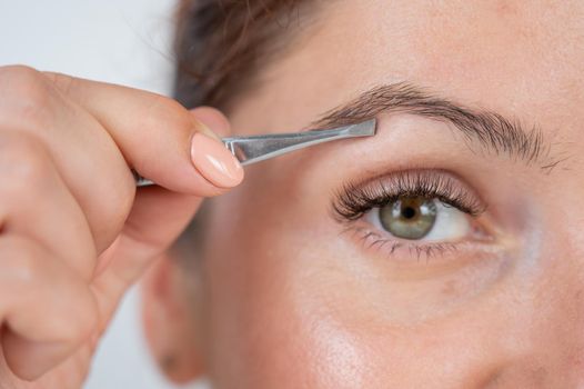 Close-up portrait of a caucasian woman doing eyebrow correction herself with tweezers