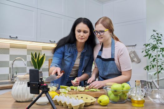 Mom and teen daughter cooking apple pie together, looking at smartphone screen