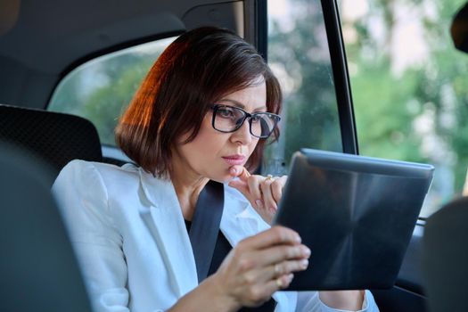 Middle-aged business woman in car in passenger backseat. Serious female in glasses with digital tablet. Business people of mature age, city, emotion fatigue meditation sadness depression frustration