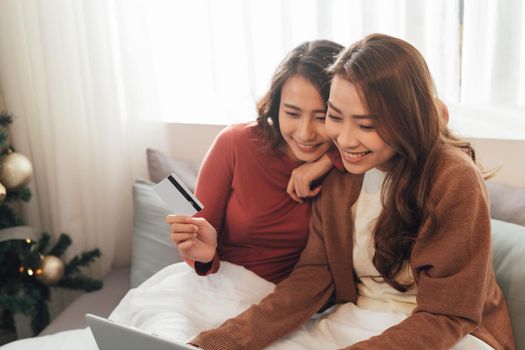 Two happy roommates buying together on line with credit card and a laptop sitting on a sofa at home with a homey background