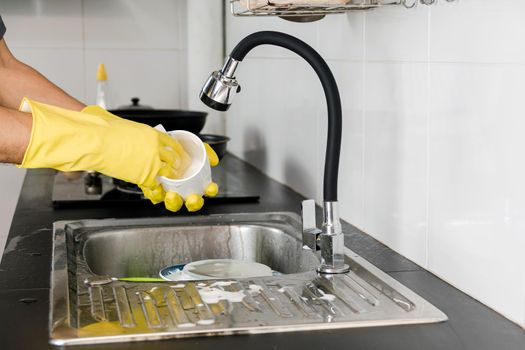 Human hands in yellow rubber gloves washing a ceramic coffee mug in the kitchen sink.