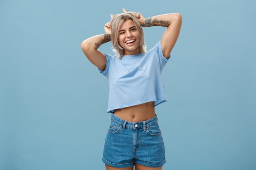Studio shot of cute attractive and joyful stylish blond woman with tattoos on arms winking and smiling broadly holding index fingers on head making horns standing in summer outfit over blue wall
