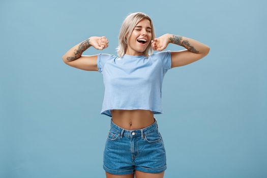 Great feelings after nap. Satisfied carefree and relaxed attractive athletic and stylish female with tattooed arms stretching hands yawning and smiling with closed eyes and pleased look after sleep