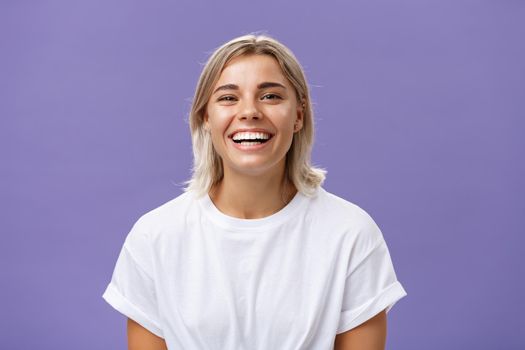 Close-up shot of joyful charming blonde female with delighted and pleased smile standing in white t-shirt over purple background spending time in awesome amusing company