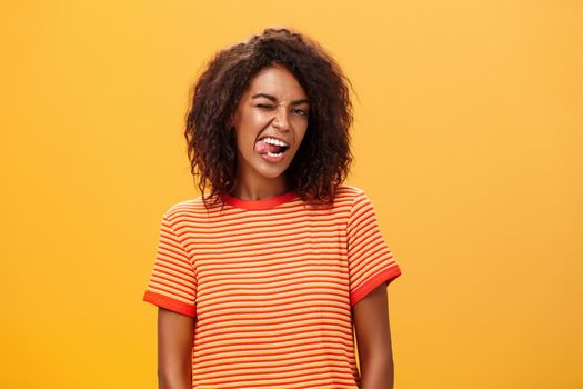 Portrait of daring and emotive confident flirty woman with afro hairstyle winking joyfully showing tongue posing carefree and enthusiastic against orange background flirting with hot guy