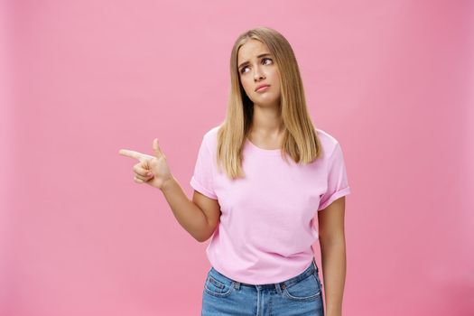 Envy upset cute young european woman with tanned skin and fair hait tilting head lifting eyebrows in sad silly look pointing, gazing left with regret and disappointment posing against pink background