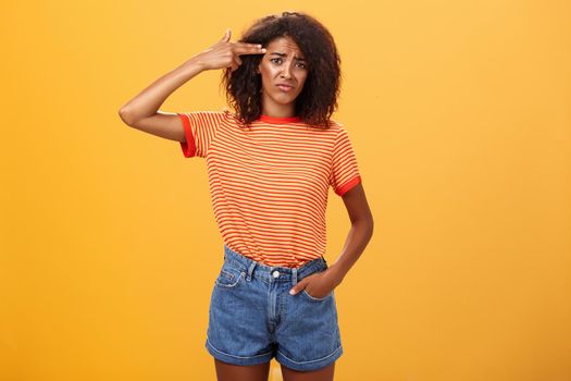 Portrait of displeased gloomy and sad annoyed dark-skinned female with curly hairstyle showing gun with hand near temple frowning wanting to kill herself from exhaust and boredom over orange wall