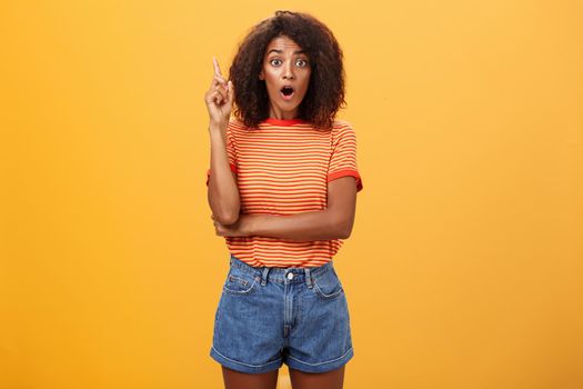 Girl finally understood riddle adding suggestion outloud. Excited thrilled good-looking dark-skinned woman staring amazed raising index finger in eureka gesture having idea over orange wall