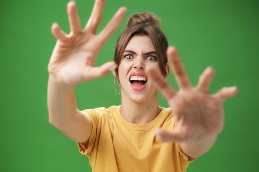 Funny emotive woman making fun face pulling hands forward to attack of grab something frowning opening mouth wanting cover face from camera, grimacing over green background