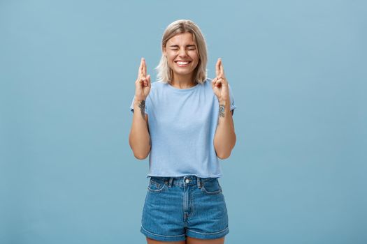 Optimistic faithful good-looking young woman with blond hair and tattoos smiling joyfully crossing fingers for good luck waiting for dream come true making wish while standing over blue background