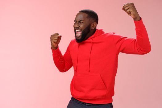 Extremely happy bearded young 25s black guy triumphing happily yelling yeah raising clenched fists celebrating success winning bet lottery standing happily pink background cheer receive great news