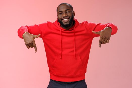Happy charismatic smiling male african american entrepreneur proudly pointing down presenting his own company product gladly showing accomplishment indicating downwards, standing pink background