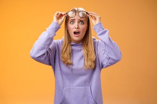 Shocked concerned young woman looking student ruin work staring disturbed upset take-off glasses popping eyes camera gasping speechless terrible acciddent happened, orange background
