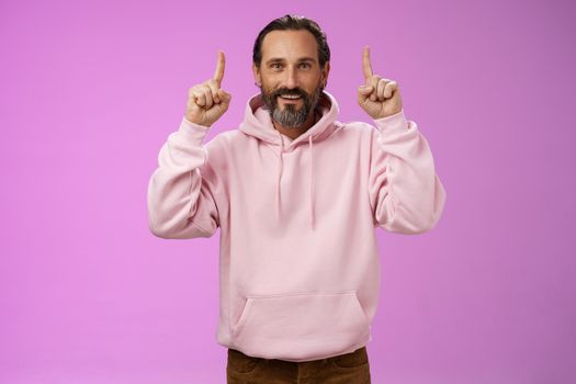 Handsome mature 50s man grey hair beard wearing stylish hipster loose hoodie pointing up index fingers showing awesome place hang out unwilling grow old standing positive lucky purple background