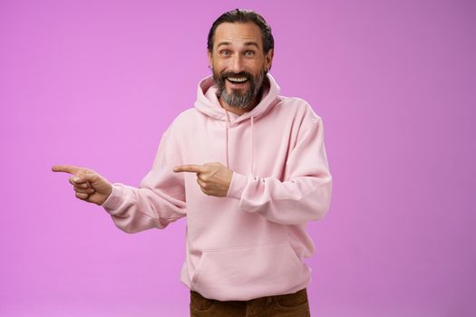 Charismatic happy good-looking mature 50s man wearing stylish hipster hoodie smiling excited pointing left index fingers impressed awesome interesting proposal standing pleased purple background