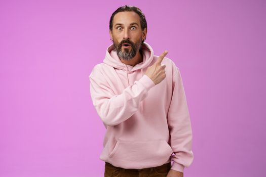 Amused handsome hipster mature 40s bearded caucasian man grey hair wearing trendy pink hoodie fold lips amazed wow pointing upper right corner widen eyes surprised check out awesome product