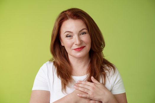 Lovely pleased tender redhead middle-aged ginger woman press hands heart feel touched grateful appreciate sincere heartwarming moment admire thankfully grin look camera glad green background