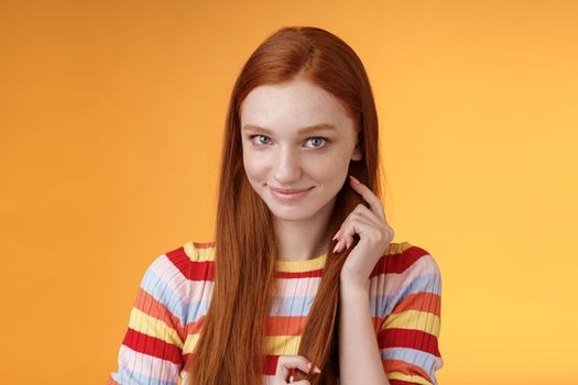 Romantic flirty shy attractive redhead girl 20s touching hair strand smiling silly modest glancing camera coquettish making lovely glances wanna seduce guy expressing sympathy, orange background