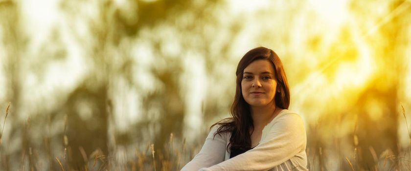 Portrait of beautiful Hispanic young woman with long hair looking at the camera against a background of unfocused trees during sunset