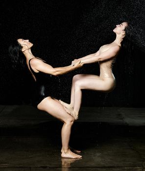 two women of Caucasian appearance perform an acrobatic stunt on a black background with rain. Woman holds on her knees another in beige bodysuit