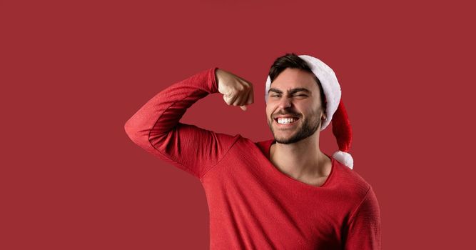 Young handsome caucasian guy in a red sweater and Santa hats stands on red background in studio and showing biceps power
