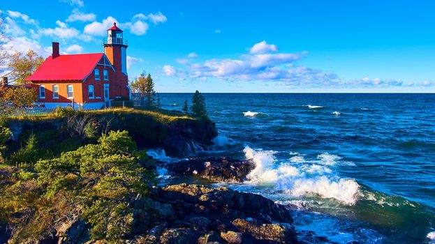 Aerial Eagle Harbor Lighthouse With crashing lake waves and green trees with a gorgeous blue sky and white and purple clouds